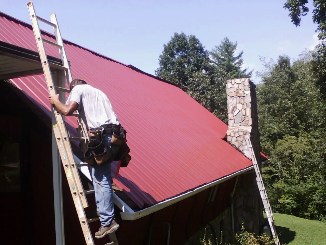 Roofer on a ladder beside a red metal roof and stone chimney — Soffit & Fascia Repair by Roofing Brothers in Newberg